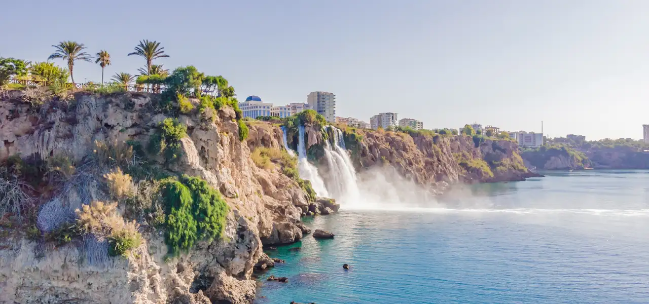 Felswand an der Küste an einem Sommertag vom Meer aus gesehen, an dem der Duden-Wasserfall in Antalya ins Meer strömt. Die Küstenlinie ist von Häusern gesäumt. 