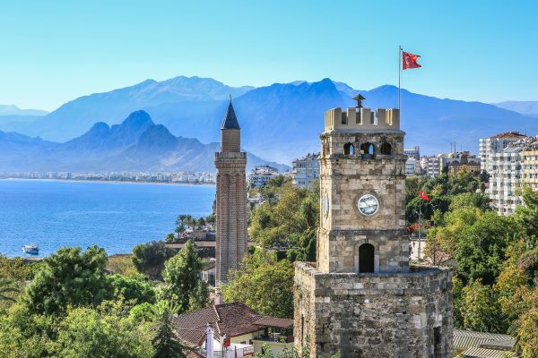 Aufnahme von der historischen Altstadt von Antalya. Im Vordergrund steht ein Turm und die Spitze eines Minarettes, umrahmt von Bäumen. Im Hintergrund sind Berge zu sehen.