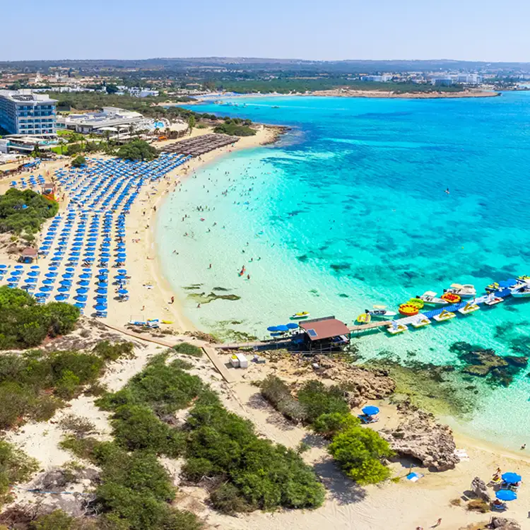 Drohnenaufnahme auf den Sandstrand in der Feigenbaumbucht an einem Sommertag. Die Küste ist mit Hotels bebaut. Am Strand stehen viele Sonnenschirme und Menschen baden im flachen, türkisfarbenem Wasser.