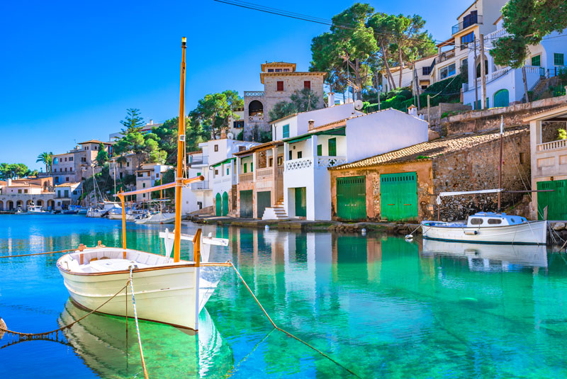 Idyllisches Fischerdorf auf der balearischen Insel Mallorca mit Wohnhäusern und kleinen Booten an einem schönen Sommertag mit strahlend blauem Himmel. 