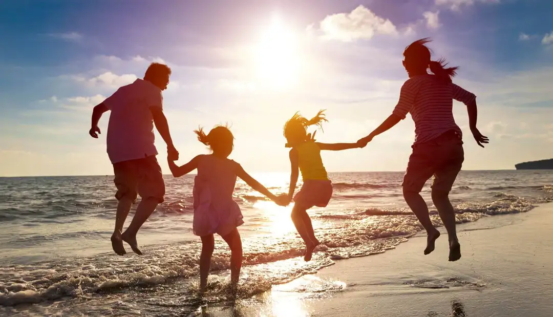 Familie mit zwei Kindern am Meer springen alle gleichzeitig in die Luft an einem sonnigen Tag. Die Sonne steht hinter den Personen, so dass nur die Silhouetten zu erkennen sind.