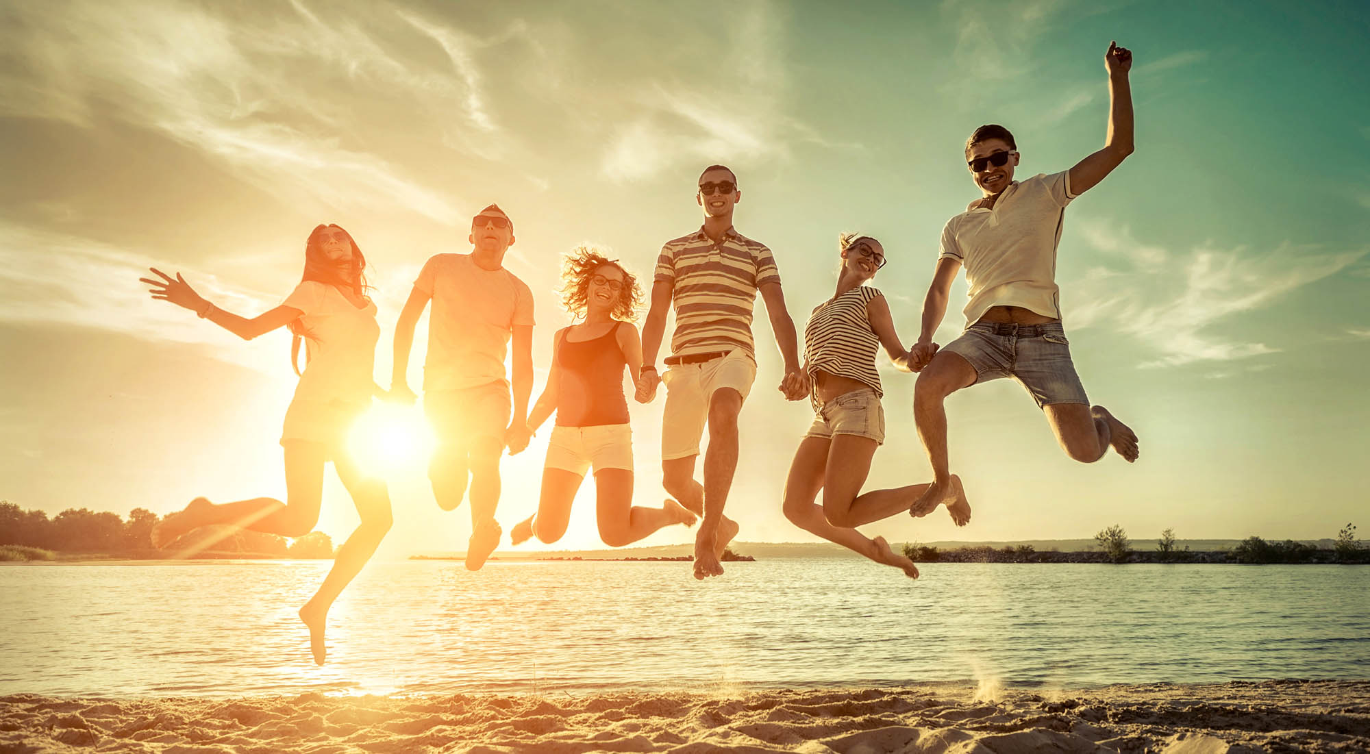 Gruppe von jungen Menschen im Urlaub in einer Reihe, die gleichzeitig in die Luft springen, vor einem See.