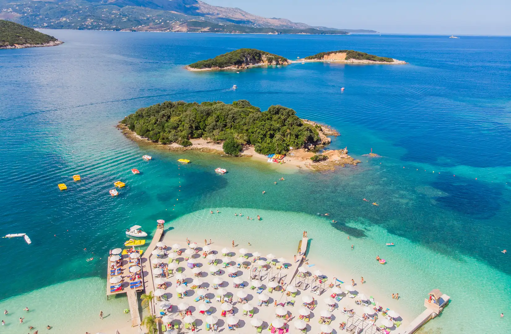 Drohnenaufnahme des Archipels der Tetraner Inseln mit den bekannten vier Inseln an einem schönen Sommertag. Im Vordergrund ist ein Strand mit Sonnenschirmen zu sehen und auf dem Wasser im Meer stehen in strandnähe zahlreiche Tretboote.