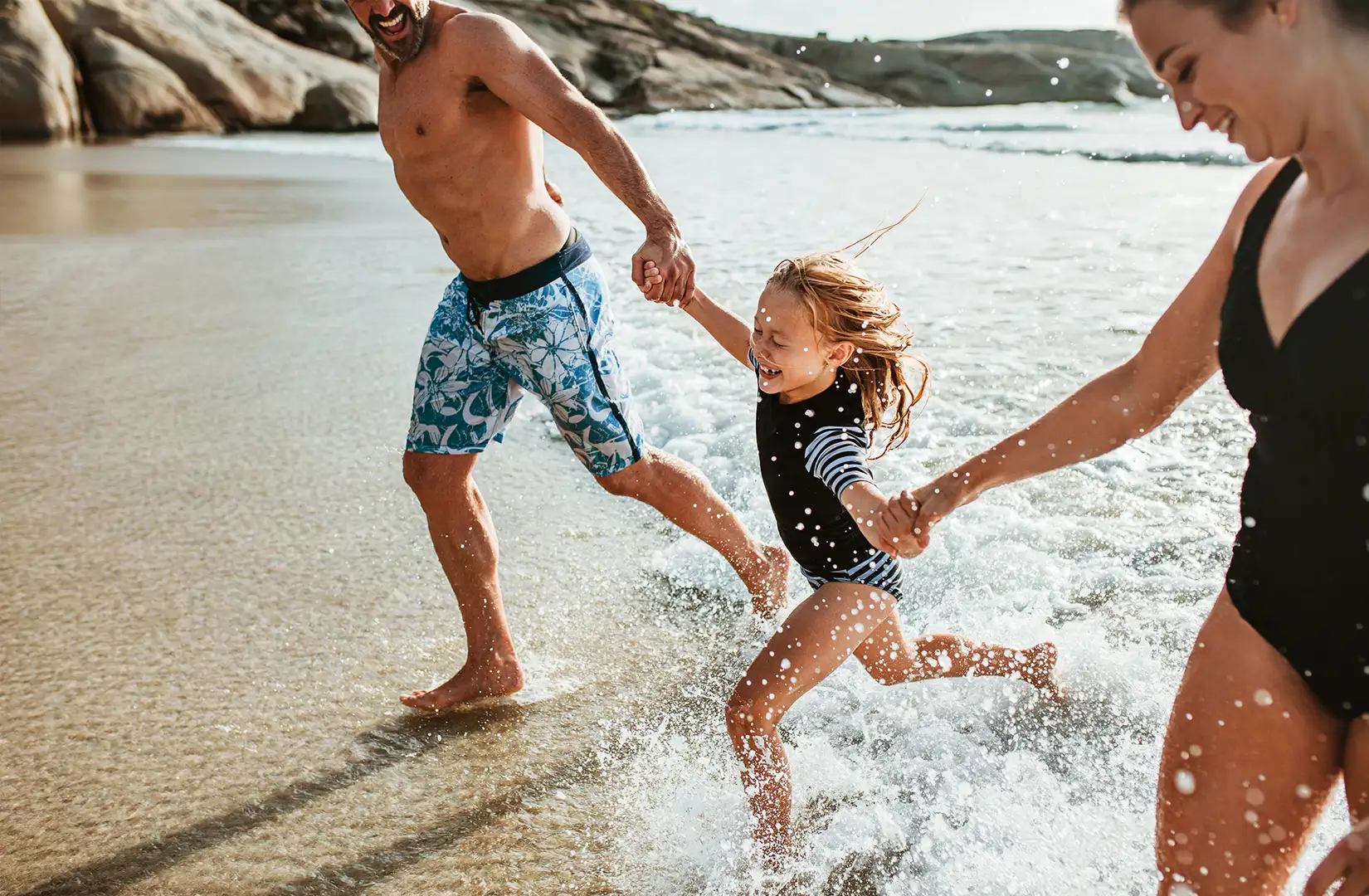 Eltern mit Kind Eltern halten ihre Tochter an den Händen und laufen am Strand aus dem Meerwasser heraus.